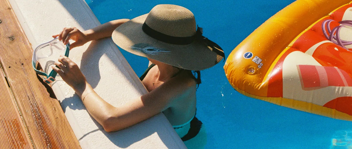 A woman in a wide-brimmed sun hat holds swim goggles while leaning on the edge of a pool, with a bright inflatable float drifting nearby