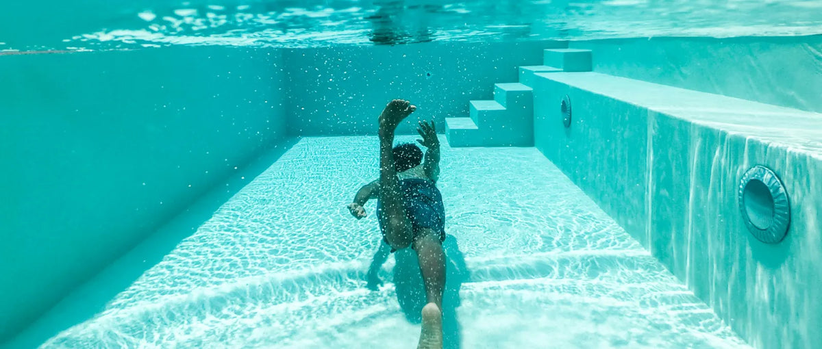 An underwater view of a person swimming in a clear, turquoise pool. Sunlight creates beautiful patterns on the floor and steps
