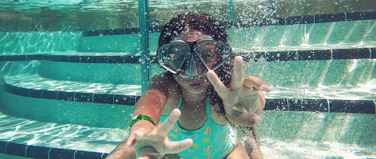 A girl wearing a snorkel mask makes peace signs while swimming underwater in a pool, surrounded by bubbles